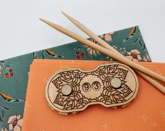May include: A wooden stitch marker with a decorative mandala design and the number "93" engraved on it. The marker is sitting on a fabric surface with two wooden knitting needles.