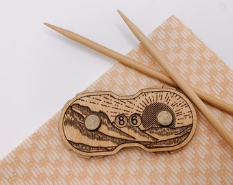 May include: A wooden stitch counter with a mountain and sun design. The counter has two brass knobs and displays the number "86".