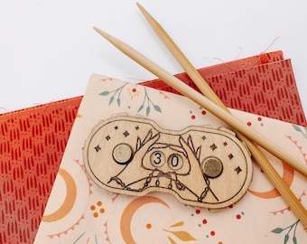 May include: Wooden stitch counter with two hands holding a chain and a number "30" in the center. The counter is on a pink and white fabric with a floral pattern. Two wooden knitting needles are on top of the fabric.