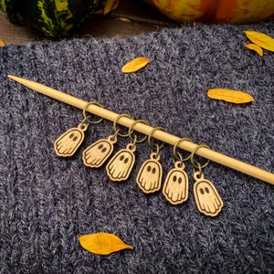 May include: Wooden ghost-shaped stitch markers on a knitting needle. The markers are attached to small metal rings. The background is a blue knitted fabric with yellow leaves, suggesting a fall or Halloween theme.