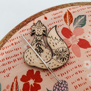 May include: A wooden fox needle minder with floral details, sitting on a pink and red floral fabric with a needle threaded through it.