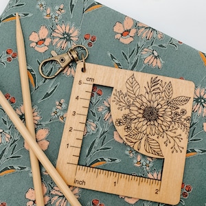 May include: Wooden ruler with a floral design, measuring up to 5 cm or 2 inches. The ruler has a key ring attached and is on a green fabric with a floral pattern.