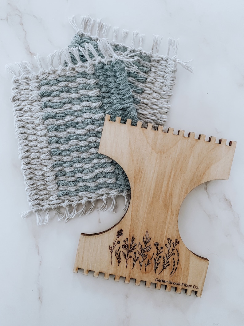 K&ouml;nnte beinhalten: Ein Holzwebstuhl mit einem Blumenmuster und dem Text "Cedar Brook Fiber Co.". Der Webstuhl wird verwendet, um einen gewebten Wandteppich mit einem blauen und wei&szlig;en Muster zu erstellen.