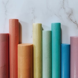 Puede incluir: Un conjunto de ocho clavijas de madera pintadas en colores del arcoíris, incluyendo rosa, naranja, amarillo, verde, azul y morado. Las clavijas están dispuestas en una fila sobre una superficie de mármol blanco.