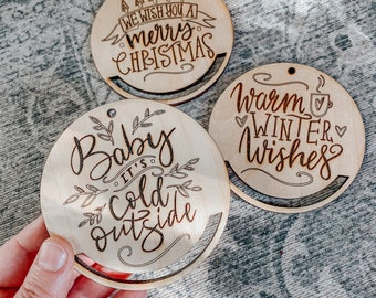 May include: Three round wooden ornaments with laser-cut designs. The first ornament says "We wish you a merry Christmas". The second ornament says "Baby it's cold outside". The third ornament says "Warm winter wishes".
