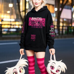 May include: Black hoodie with pink graphic text and design. The text includes "ANGEL" and "DADDY'S LITTLE SINNER". The person is wearing pink and white striped knee-high socks and pink boots. Two white plush bags with cartoon faces are held.