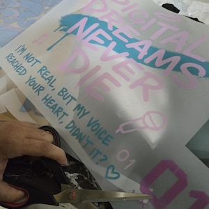 K&ouml;nnte beinhalten: Ein Bogen wei&szlig;es Transferpapier mit dem Text "DIGITAL DREAMS NEVER DIE" in Rosa, mit einem blauen Spr&uuml;heffekt. Der Text "I'M NOT REAL, BUT MY VOICE REACHED YOUR HEART, DIDN'T IT?" ist ebenfalls sichtbar. Eine Schere schneidet das Papier.