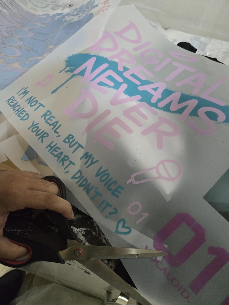 K&ouml;nnte beinhalten: Ein Bogen wei&szlig;es Transferpapier mit dem Text "DIGITAL DREAMS NEVER DIE" in Rosa, mit einem blauen Spr&uuml;heffekt. Der Text "I'M NOT REAL, BUT MY VOICE REACHED YOUR HEART, DIDN'T IT?" ist ebenfalls sichtbar. Eine Schere schneidet das Papier.