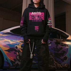 May include: Black hoodie with the word "ANGEL" in a pink box, featuring a graphic design. The hoodie also has text that reads "DADDY'S LITTLE SINNER." The person is wearing black pants and a silver chain necklace. A car with colorful graphics is in the background.
