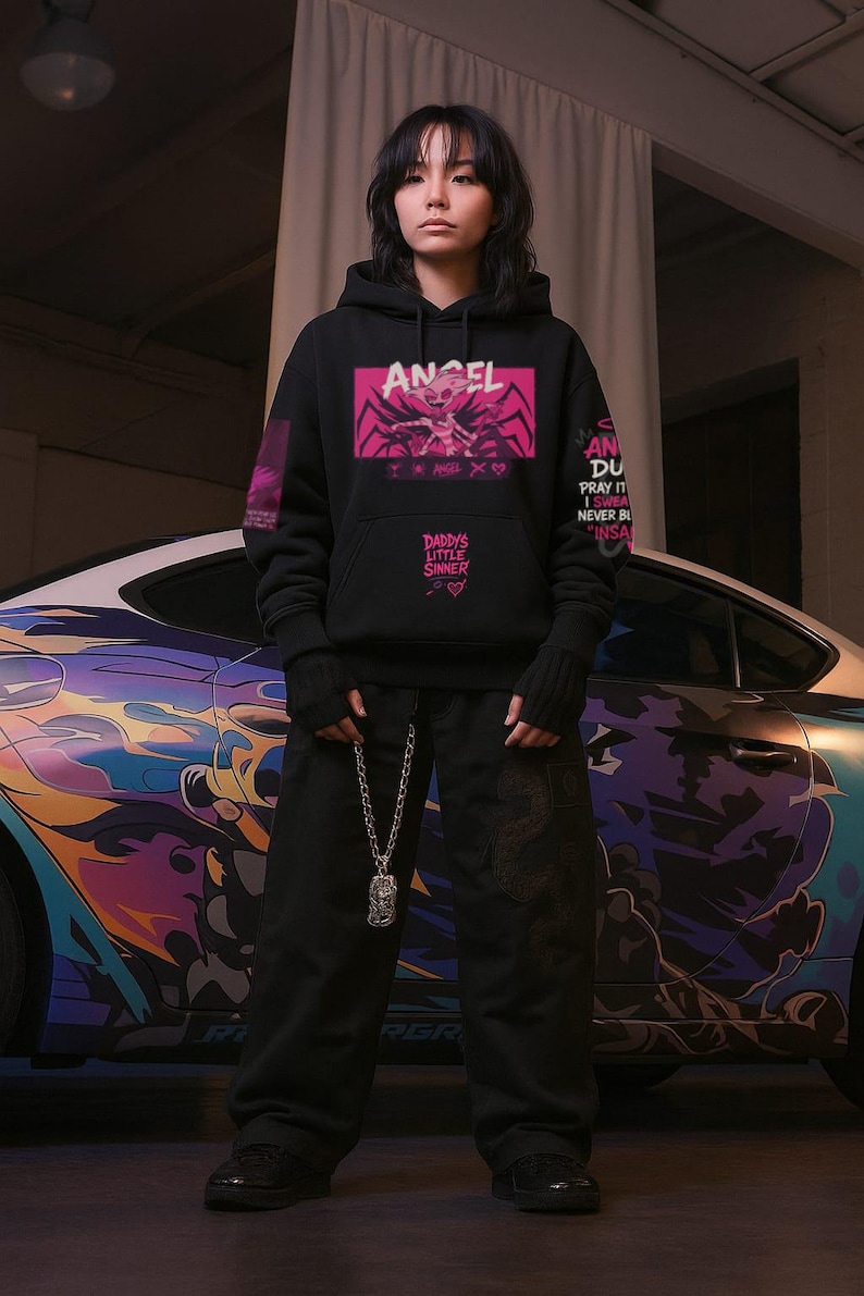 May include: Black hoodie with the word "ANGEL" in a pink box, featuring a graphic design. The hoodie also has text that reads "DADDY'S LITTLE SINNER." The person is wearing black pants and a silver chain necklace. A car with colorful graphics is in the background.