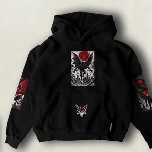 Può includere: Felpa con cappuccio nera con una grafica dettagliata di una creatura simile a un pipistrello con una rosa rossa e un design di paesaggio urbano. Le maniche presentano opere d'arte simili. Un piccolo logo è sulla tasca anteriore e la parola "DESIGN" è stampata su un'etichetta nera.