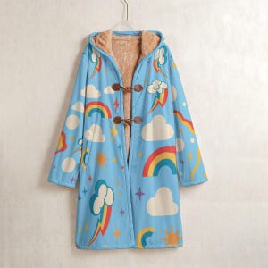 Op de afbeelding: Een lichtblauwe jas met capuchon en een kleurrijk regenboog- en wolkenpatroon. De jas heeft een bruine imitatiebontvoering en houten knevelsluitingen. Het ontwerp omvat regenbogen, wolken, sterren en bliksem.