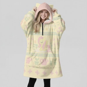 May include: A light yellow hooded blanket with a pink cat ear hood and a black zipper. The blanket features a cartoon character design with flowers and watering cans. The design is in pastel colors.