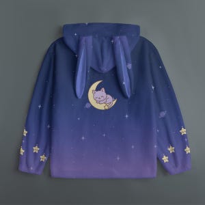 Op de afbeelding: Een hoodie met blauwe en paarse kleurovergang, met een grillig ontwerp. De capuchon heeft bevestigde konijnenoren. De achterkant toont een slapende kat op een maan, omringd door sterren en planeten. Gele steraccenten sieren de mouwen.