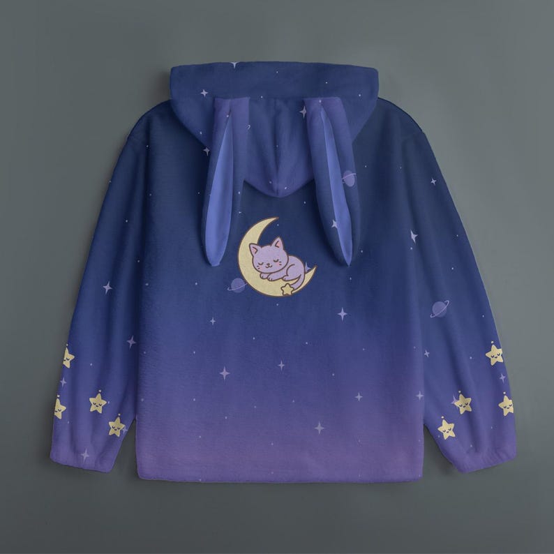 Op de afbeelding: Een hoodie met blauwe en paarse kleurovergang, met een grillig ontwerp. De capuchon heeft bevestigde konijnenoren. De achterkant toont een slapende kat op een maan, omringd door sterren en planeten. Gele steraccenten sieren de mouwen.