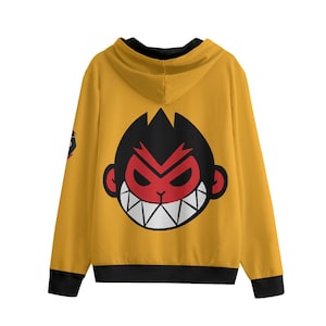 Op de afbeelding: Mosterdgele hoodie met een groot cartoon apengezicht op de rug. De aap heeft een rood gezicht, zwart haar en witte tanden. De hoodie heeft zwarte afwerking op de manchetten en zoom.