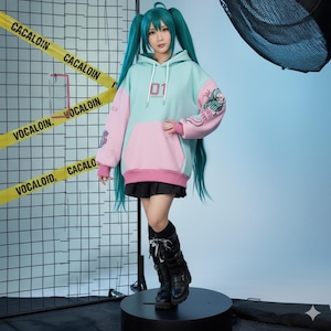 K&ouml;nnte beinhalten: Ein hellblauer und rosa Kapuzenpullover mit der Nummer "01" auf der Vorderseite. Der Hoodie hat eine K&auml;ngurutasche und eine Grafik auf dem &Auml;rmel. Die Person tr&auml;gt einen schwarzen Rock und schwarze Stiefel. Gelbes Warnband mit dem Wort "CACALOIN" im Hintergrund.