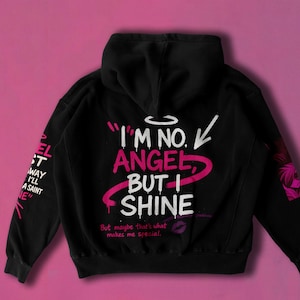 May include: Black hoodie with the text "I'M NO. ANGEL, BUT I SHINE" in white and pink. The left sleeve has the text "EL ST WAY I'LL A SAINT NE" in white. A pink background complements the design.