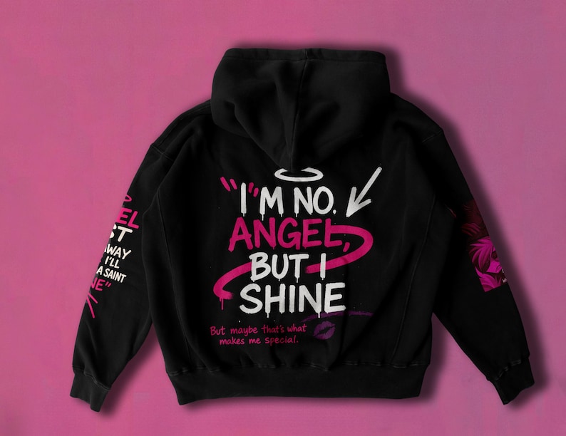May include: Black hoodie with the text "I'M NO. ANGEL, BUT I SHINE" in white and pink. The left sleeve has the text "EL ST WAY I'LL A SAINT NE" in white. A pink background complements the design.
