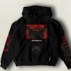Cryptid-themed Mothman hoodie featuring the mysterious winged figure with glowing red eyes, silhouetted against a dark sky or bridge scene, perfect for fans of paranormal legends and eerie aesthetics.
