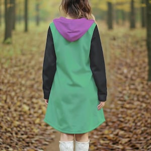 May include: A green hooded dress with black sleeves and a purple hood. The dress is knee-length and has a relaxed fit. The background is a blurred forest scene with fallen leaves.