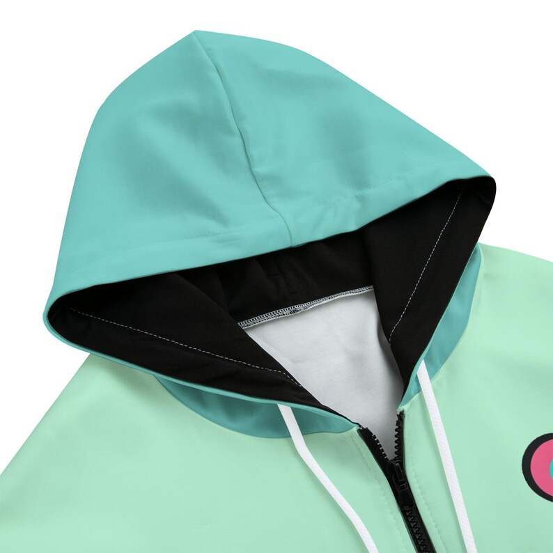 May include: Close-up of a mint green and teal hoodie with a black interior lining. The hood is up, and the zipper is partially closed. White drawstrings are visible. A pink and white graphic is on the lower right.