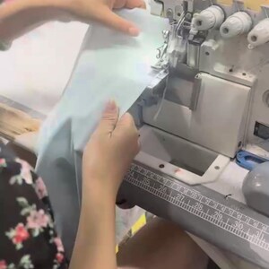 May include: A person operating a Siruba 647KP sewing machine. The machine is silver and gray, with a ruler along the front. The person is holding light blue fabric, preparing to sew it.
