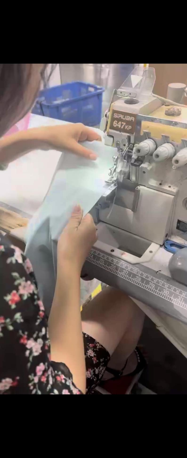 May include: A person operating a Siruba 647KP sewing machine. The machine is silver and gray, with a ruler along the front. The person is holding light blue fabric, preparing to sew it.