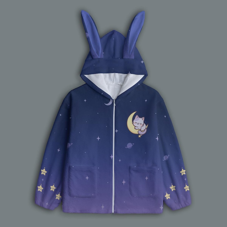 Op de afbeelding: Een marineblauwe hoodie met ritssluiting en een sterrenhemelontwerp, met een kat die op een maan slaapt. De hoodie heeft een capuchon met konijnenoren, twee voorzakken en steraccenten.