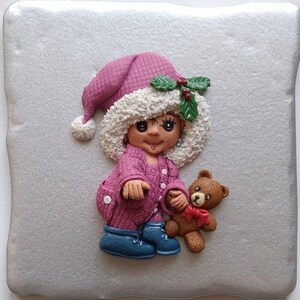 Puede incluir: Una figurita rosa y blanca de un niño que lleva un gorro de Papá Noel y sostiene un oso de peluche marrón. El niño lleva un suéter rosa y botas azules. La figurita está sobre un fondo plateado.