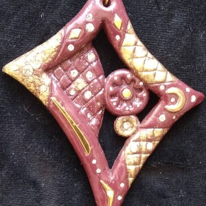 May include: A burgundy diamond-shaped ornament with gold accents and white rhinestones. The ornament features a geometric design with a circular pattern in the center.