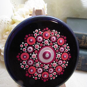 May include: A dark blue, circular decorative item with a mandala design. It features concentric circles in red, pink, and white. The item is displayed on a small wooden easel.