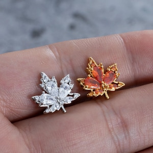 May include: Two leaf-shaped earrings. One is silver-toned with clear, sparkling stones. The other is gold-toned with orange-red stones. Both earrings are detailed with a maple leaf design, perfect for autumn or nature-themed jewellery.