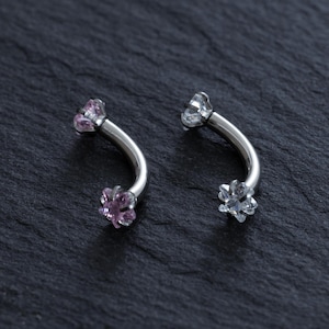 Pode incluir: Duas peças de joalharia corporal em forma de barra curva feitas de metal prateado polido. Cada barra apresenta uma pequena pedra preciosa facetada em forma de estrela em ambas as extremidades. Uma peça tem pedras rosa claro, a outra pedras incolores.
