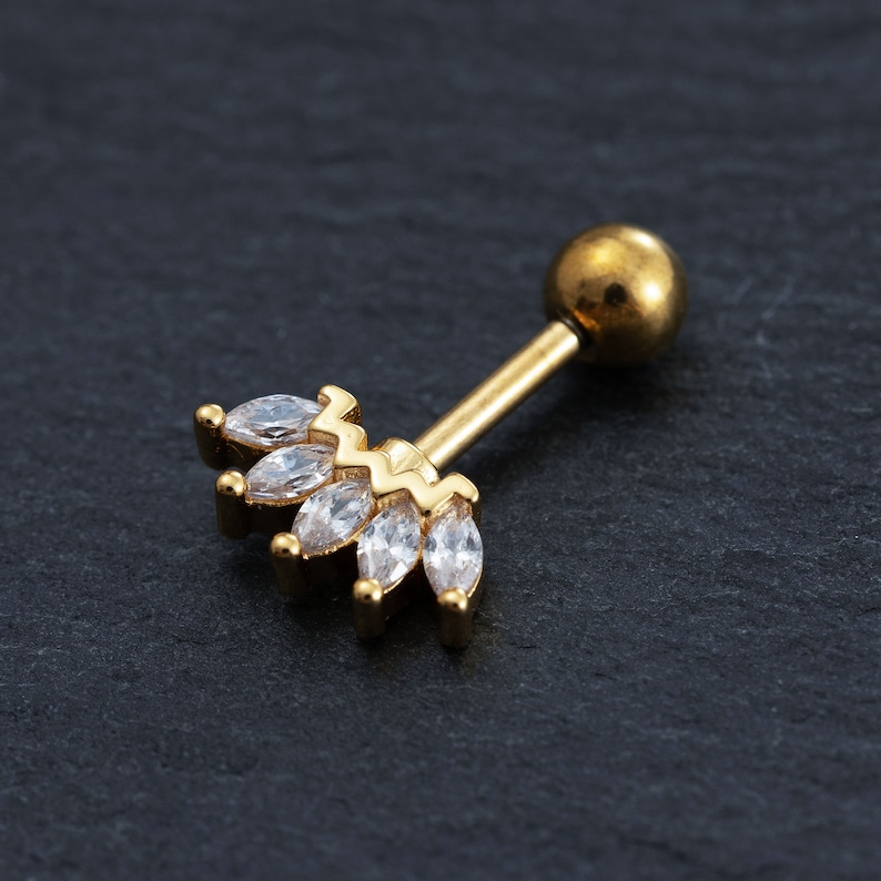 May include: Gold body jewellery with a clear crystal crown design. The jewellery is a barbell style with a round ball on one end and a crown of clear crystals on the other end.