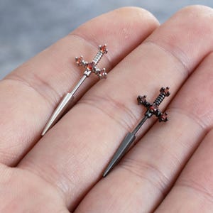 Può includere: Due gioielli per il corpo a forma di spada, uno in metallo color argento e uno in metallo nero. Ognuno presenta una lama appuntita, una guardia e un'impugnatura decorata con piccole pietre preziose rosse rotonde incastonate nel metallo.
