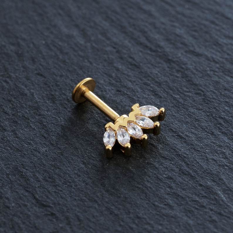 May include: Gold body jewellery with a clear marquise-shaped stone cluster. The cluster is in the shape of a crown.