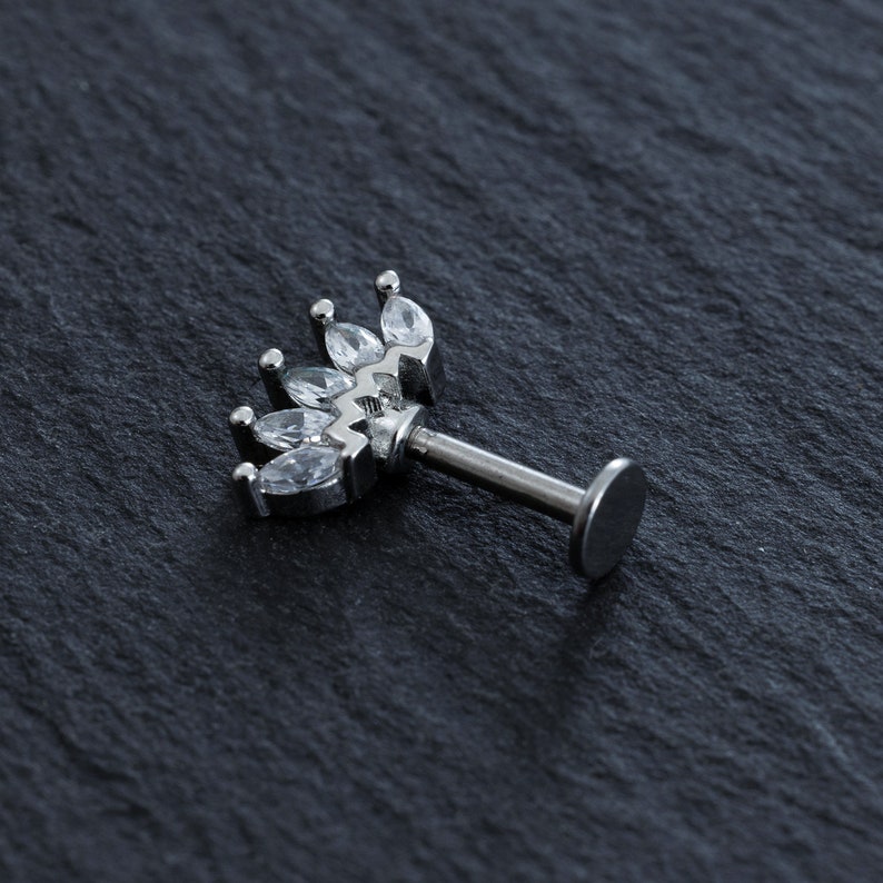May include: Silver body piercing with a clear crystal crown design. The crown is made of small, clear, marquise-shaped stones.
