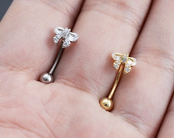 May include: Two belly button rings with a bow design. The bow is adorned with clear cubic zirconia stones. One ring is silver and the other is gold.