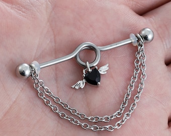Puede incluir: Un piercing industrial de color plateado con un dije en forma de corazón negro con pequeñas alas y dos cadenas colgantes. La barra tiene un diseño curvo y dos extremos de bola redonda.