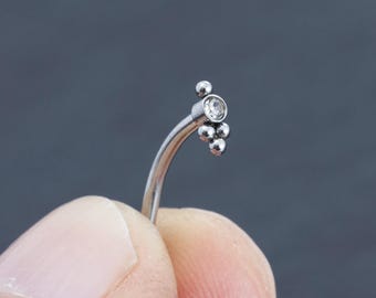 May include: A curved, silver-colored body jewelry piece. It features a cluster of small, round beads and a single, larger, clear gemstone. The jewelry is held against a gray background.