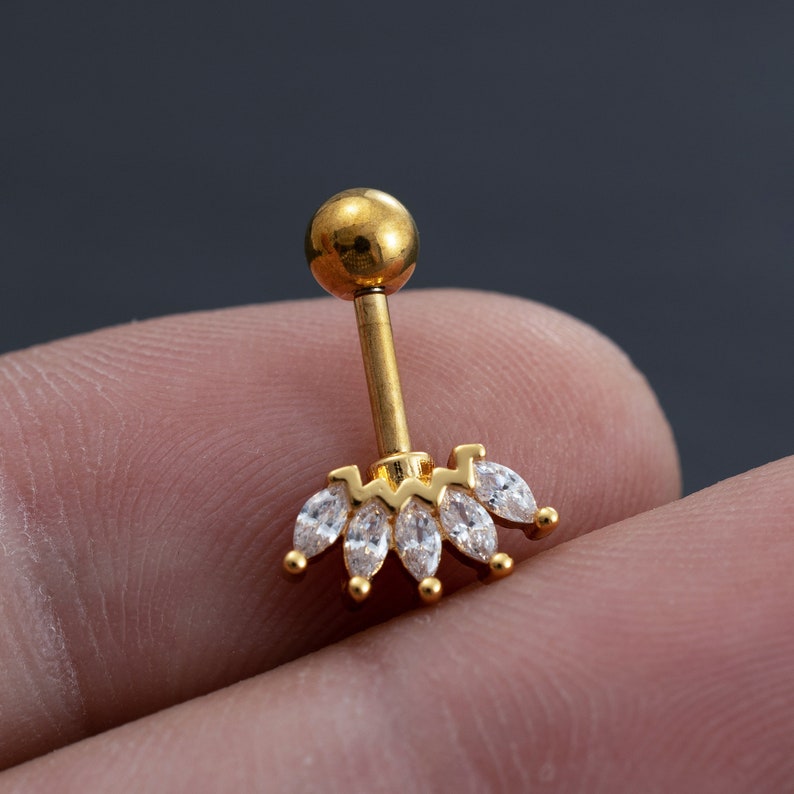 May include: Gold body jewellery with a clear crystal marquise shaped stone cluster. The jewellery is a barbell style with a round ball on the top.