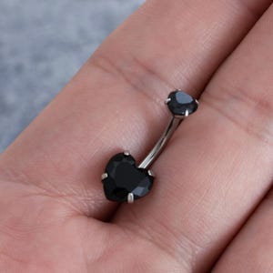 Könnte beinhalten: Ein silberner Bauchnabelring mit zwei schwarzen herzförmigen Steinen.