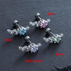 May include: Four stud earrings with a star-shaped center stone in blue, pink, light purple, and green. Each earring has a silver post and is surrounded by small, clear crystals. The earrings are displayed on a dark gray surface.