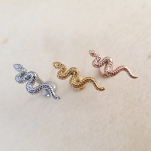 May include: Three snake-shaped body jewellery pieces in silver, gold, and rose gold. The jewellery is designed to be worn as a piercing.