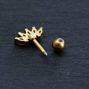 May include: Gold body jewellery with a crown-shaped top and a round ball. The jewellery is on a black surface.