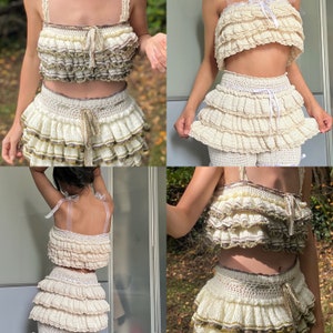 May include: A white crocheted two-piece outfit with a ruffled top and skirt. The top has a brown trim and a white ribbon tie. The skirt has multiple layers of ruffles and a brown trim.