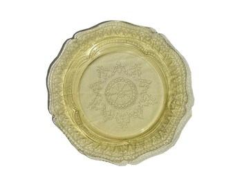 May include: A vintage yellow glass plate with a scalloped edge and intricate embossed floral and geometric patterns. The plate features a central design and a detailed border, showcasing a classic, elegant aesthetic. The plate is likely used for serving or display.