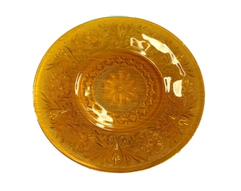 May include: A round, amber-colored glass plate with a floral and swirl pattern. The plate has a raised, textured design with a central flower motif and a wide rim. The plate is made of a transparent glass material.