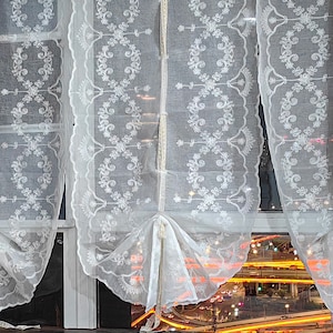 Könnte beinhalten: Weiße Spitzenvorhänge mit einem zarten Blumenmuster hängen an einem Fenster, durch den durchscheinenden Stoff ist ein verschwommener Blick auf die Lichter der Stadt und den Verkehr zu sehen.